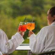 Twee dames in badjas proosten met cocktail met uitzicht over de natuur.